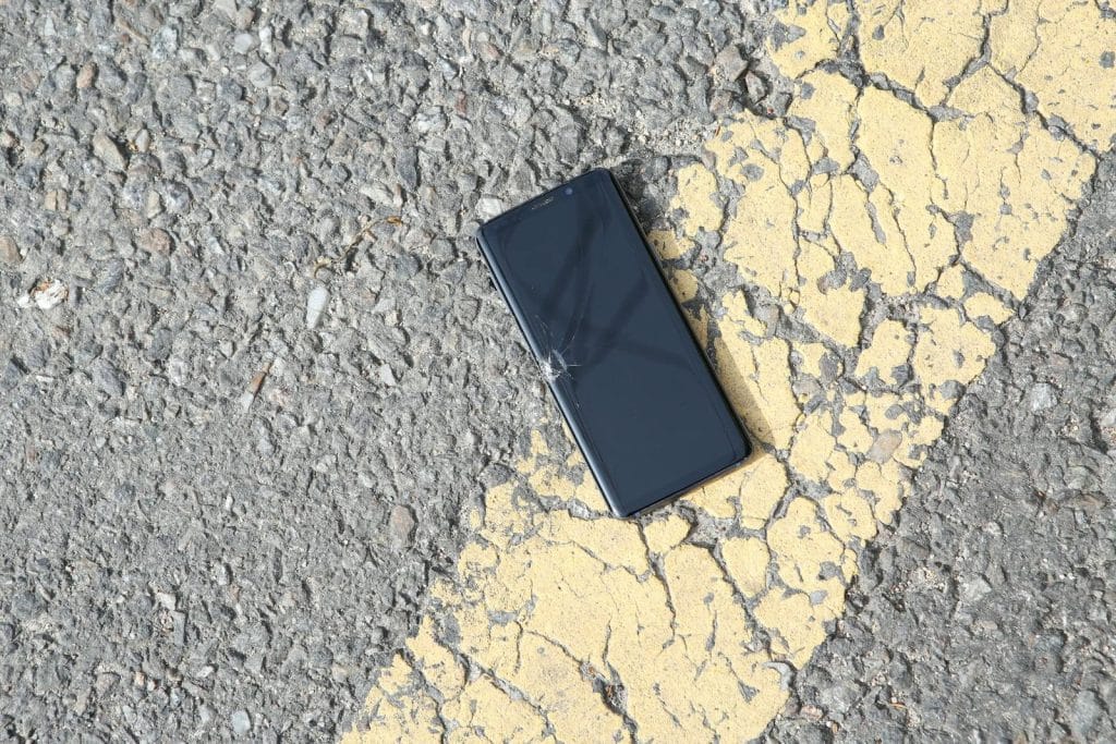 A close-up view of a cracked smartphone lying on a sunlit asphalt road.